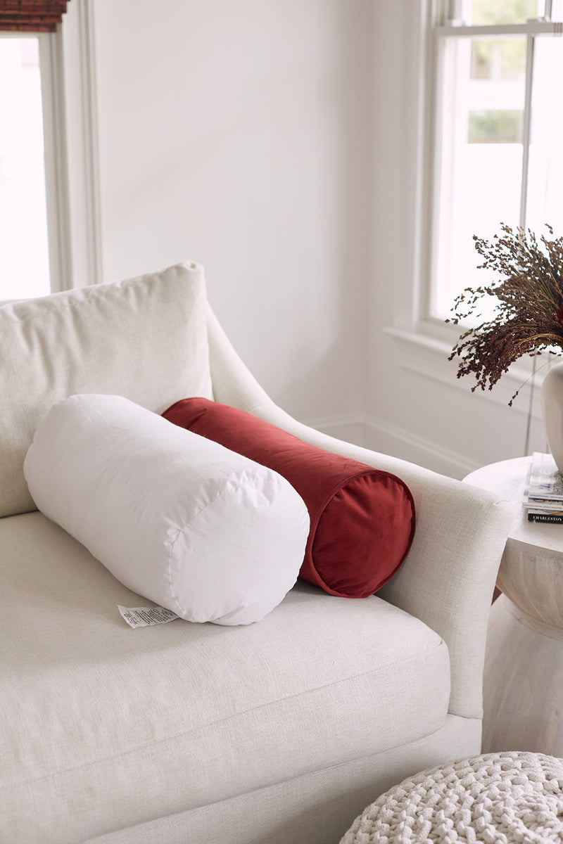 Two Sewing Down South 8 x 20 Bolster Indoor Inserts, one in white and one in rust, are placed side by side on a light cream sofa in a sunlit room. A dried floral arrangement sits on a nearby round table.