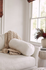 A cozy beige sofa with a chunky knit blanket and a white cylindrical pillow featuring the Sewing Down South 8 x 20 Bolster Indoor Insert sits by a window. A side table holds dried flowers and books, creating an inviting atmosphere.