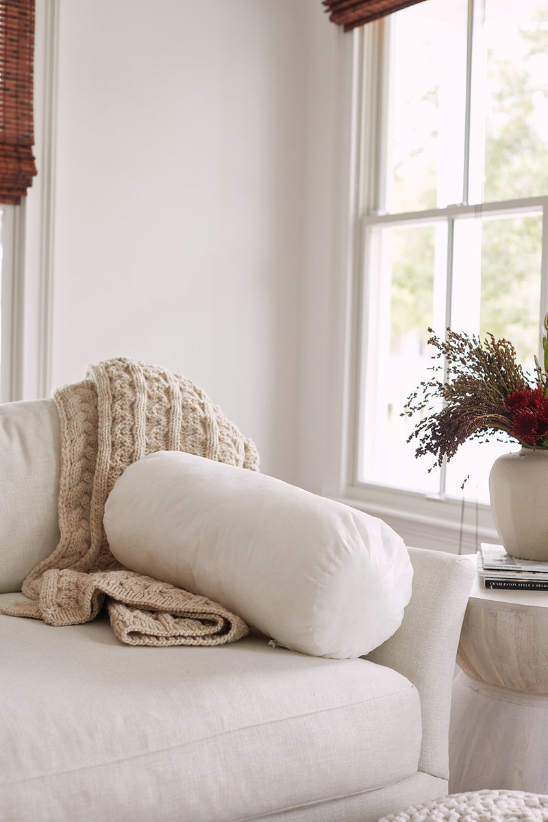 A cozy beige sofa with a chunky knit blanket and a white cylindrical pillow featuring the Sewing Down South 8 x 20 Bolster Indoor Insert sits by a window. A side table holds dried flowers and books, creating an inviting atmosphere.