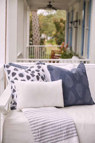 A porch swing with a striped blanket and three decorative pillows—including one with the Sewing Down South 14 x 24 Outdoor Insert—rests on a covered porch with trees and chairs in the background.