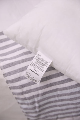 A close-up of the Sewing Down South 14 x 24 Outdoor Insert with its care label, resting on white and gray striped bedding. The label provides washing instructions for its mold resistant polyester cluster fiber fill.