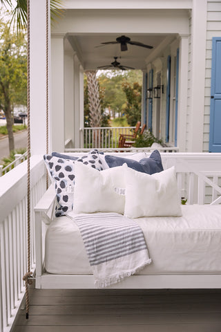 A white porch swing with blue and white pillows featuring the Sewing Down South 16 x 20 Outdoor Insert and a striped throw blanket sits on a covered porch with white railings, trees, and houses in the background.