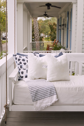 A white porch swing with mold-resistant pillows featuring the Sewing Down South 20 x 20 Outdoor Insert and a striped throw blanket is on a porch with a ceiling fan, railing, and trees in the background.