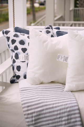 A porch swing with Sewing Down South 20 x 20 Outdoor Inserts and a folded gray striped blanket creates a cozy, inviting outdoor seating area. Soft, mold-resistant pillows feature visible tags.