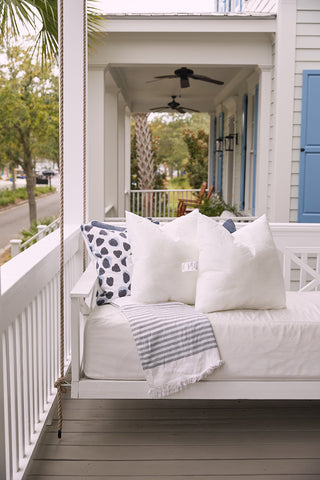 A cozy porch swing bed with white and patterned cushions featuring the Sewing Down South 23 x 23 Indoor Inserts and a striped blanket graces a wooden porch with railing. Blue shutters, a ceiling fan, and trees enhance the houses serene charm.