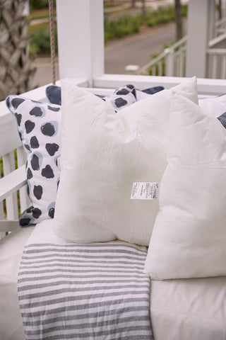 White pillows and a polka dot pillow, paired with the Sewing Down South 23 x 23 Outdoor Insert, decorate the porch swing. A striped blanket is draped over the cushions. In the background, a white railing reveals a glimpse of a tree and street.