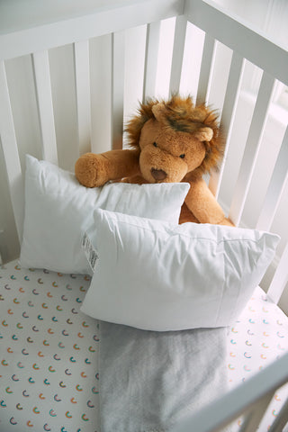 White slatted crib with a lion plush toy, two Sewing Down South 13 x 17 Indoor Nursery Inserts pillows, and a fitted sheet with small colorful rainbows. A light gray blanket is folded at the foot of the crib.