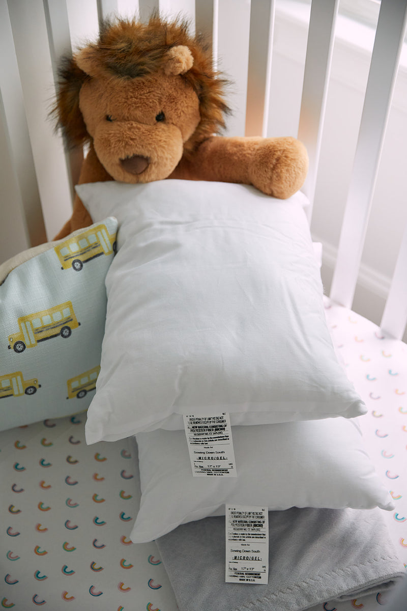 A plush lion toy rests on two white Sewing Down South 13 x 17 Indoor Nursery Inserts in a crib. Nearby, a cushion features yellow school bus illustrations, and the crib sheet has colorful rainbows. Made with non-allergenic materials, it ensures a safe, cozy space for your little one.