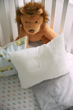 A stuffed lion toy sits in a crib with white slatted sides. Cozy nursery pillows, featuring a yellow school bus pattern, are in front of the lion. The Sewing Down South 13 x 17 Indoor Nursery Inserts provide plush comfort, and the bedding showcases a subtle rainbow design on a light background.