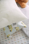 A white pillow is placed on a yellow school bus-patterned pillow, perfect for nursery decor, with the fabric tag visible. In the background, a bedspread with rainbow arches accents the scene as part of a stuffed animal peeks in. Product: Sewing Down South 13 x 17 Indoor Nursery Inserts.