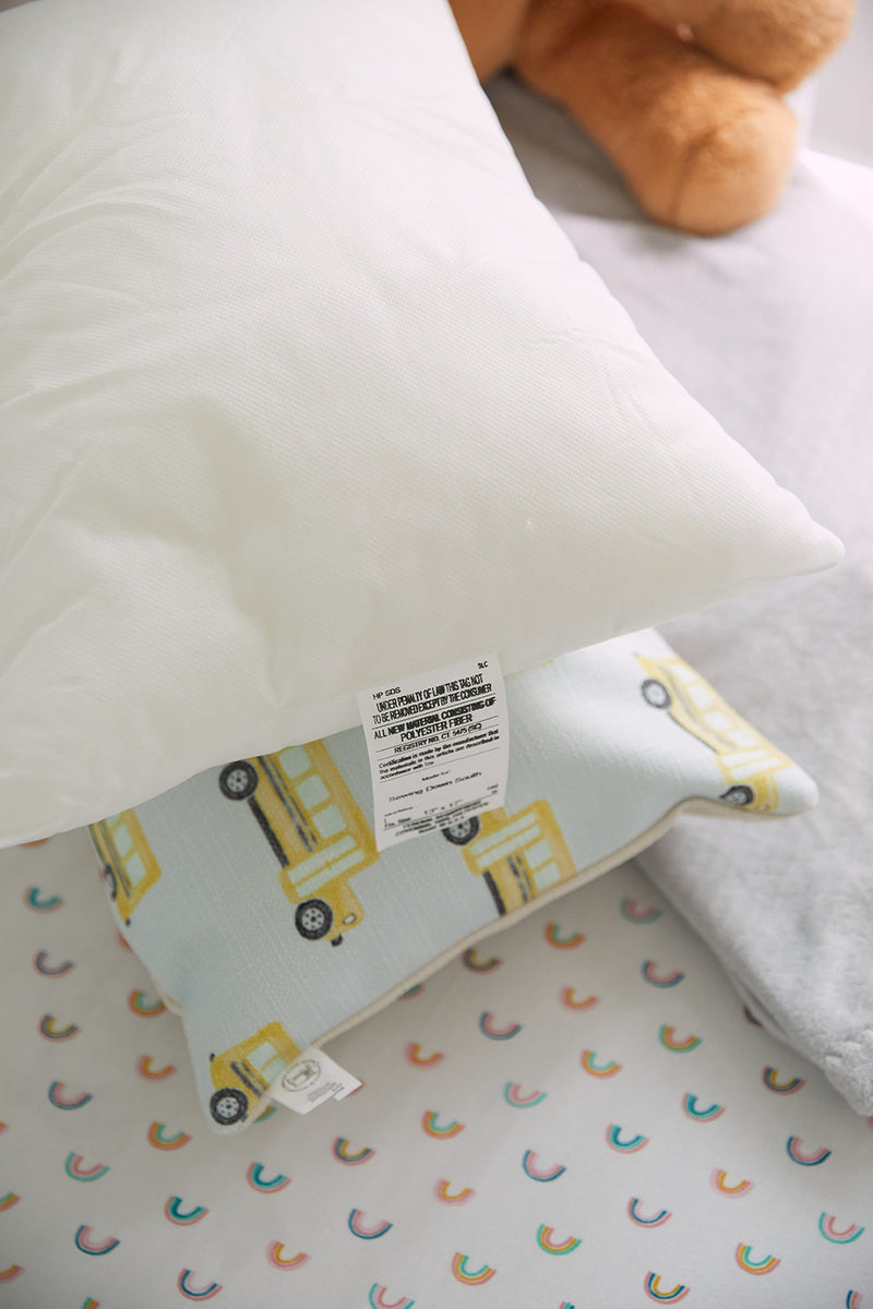 A white pillow is placed on a yellow school bus-patterned pillow, perfect for nursery decor, with the fabric tag visible. In the background, a bedspread with rainbow arches accents the scene as part of a stuffed animal peeks in. Product: Sewing Down South 13 x 17 Indoor Nursery Inserts.