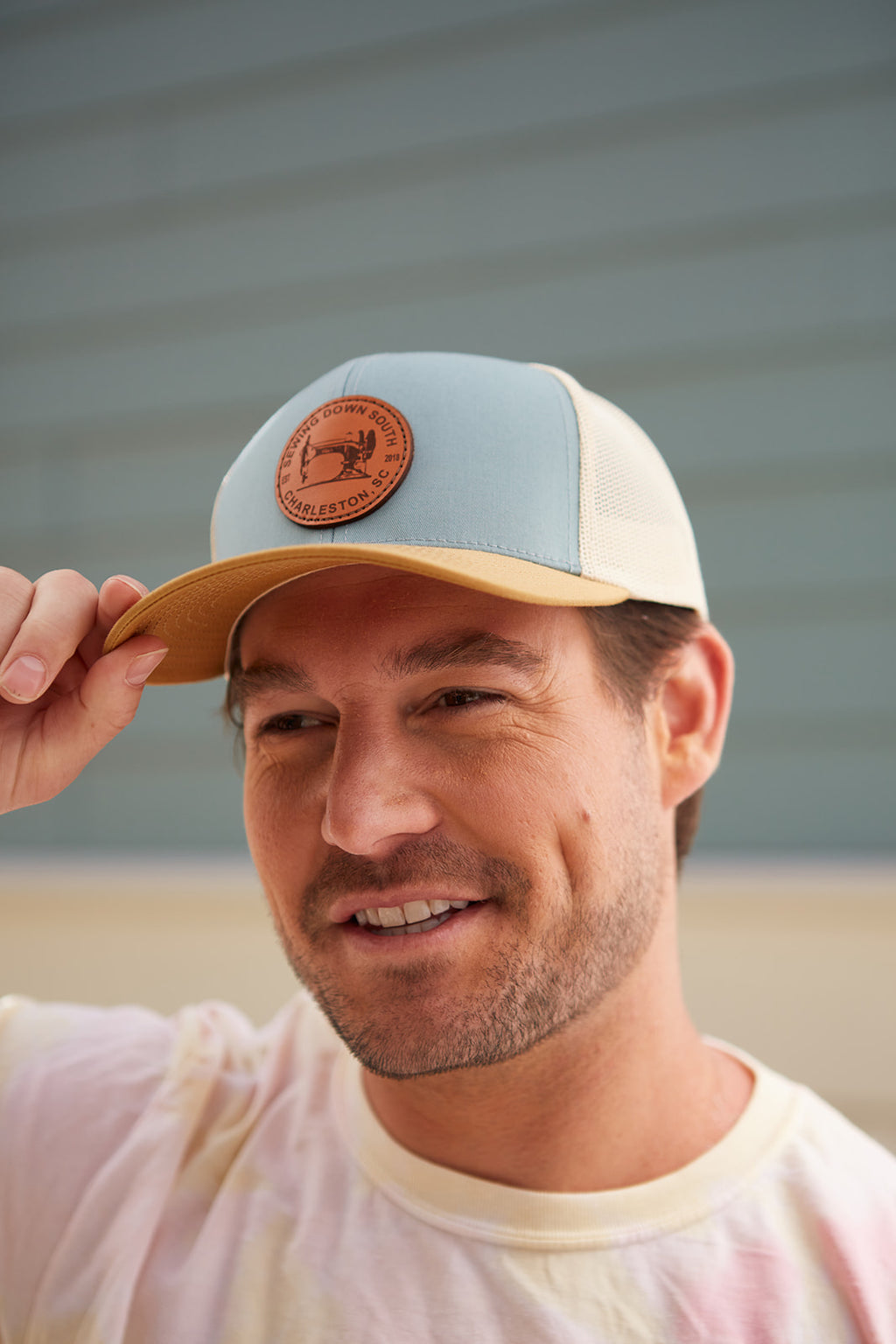 A man in a Sewing Down South Leather Patch Trucker in Smoke Blue smiles, touching the brim, with a softly blurred background highlighting his relaxed demeanor.