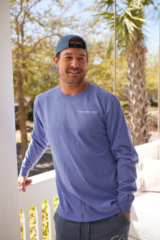 A man in a Sewing Down South Long-Sleeve Logo Tee, Rainbow Row Colors, smiles while standing on a porch with white railing, surrounded by trees and a palm. The sunny day scene complements the vintage vibe.