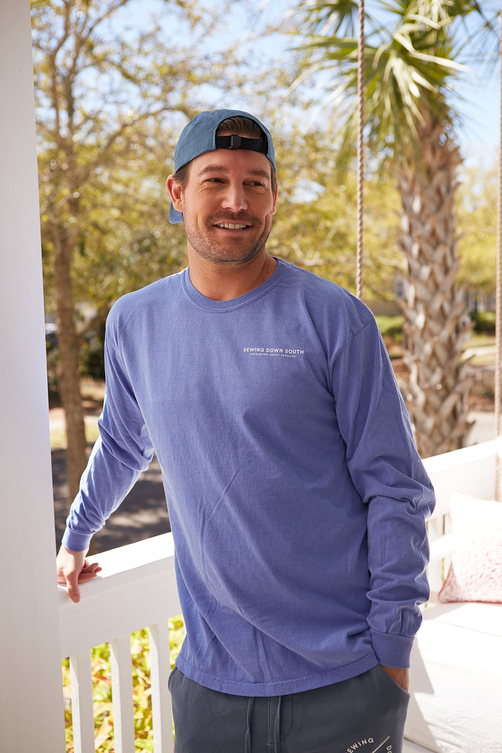 A man in a Sewing Down South Long-Sleeve Logo Tee, Rainbow Row Colors, smiles while standing on a porch with white railing, surrounded by trees and a palm. The sunny day scene complements the vintage vibe.