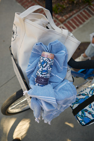 A can in a Crab Craze Koozie, 4 PACK (Skinny or Standard), featuring colorful abstract designs, rests on a blue and white towel inside a white tote bag in a bicycle basket, with part of the bike and brick sidewalk visible.