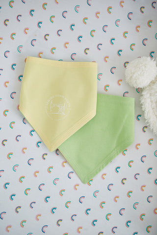 Two SDS Bandana Bibs made of cotton rest on a patterned surface with small rainbow arches; one reversible bib is yellow with white text, and the other is green. A fluffy white stuffed toy peeks from the right, making it a charming baby gift for any occasion.