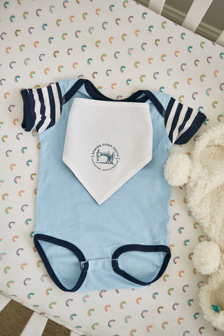 A light blue baby onesie with navy trim and striped sleeves sits on a crib mattress with a rainbow-patterned sheet. On top is the SDS Bandana Bib, a white cotton bib featuring a small circular logo with text and an illustration of a bed. Nearby, a plush toy completes this perfect baby gift set.