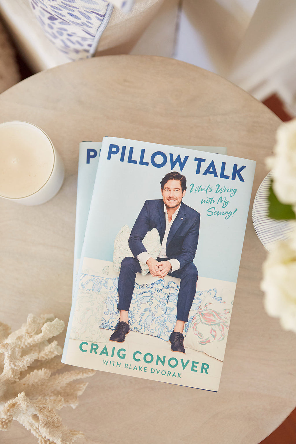 Two Exclusive: Signed Pillow Talk Book copies by Southern Charms Craig Conover and Blake Dvorak, featuring a suited man on the cover, rest on a table. A flickering white candle completes the perfect Southern evening ambiance.