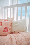 A cozy crib with a white railing holds a pink blanket and two pillows: an Organic Toddler Sleeping Pillow and another pink one with rainbow designs. Sunlight streams through a nearby window, casting gentle beams across this charming setup.