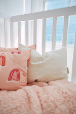 A cozy crib with a white railing holds a pink blanket and two pillows: an Organic Toddler Sleeping Pillow and another pink one with rainbow designs. Sunlight streams through a nearby window, casting gentle beams across this charming setup.