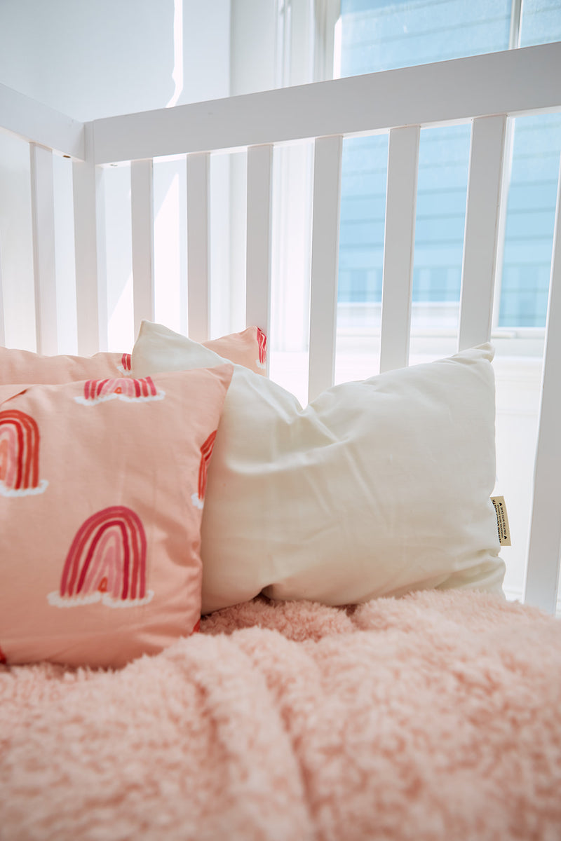 A cozy crib with a white railing holds a pink blanket and two pillows: an Organic Toddler Sleeping Pillow and another pink one with rainbow designs. Sunlight streams through a nearby window, casting gentle beams across this charming setup.