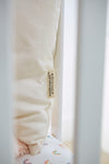 A close-up of the Organic Toddler Sleeping Pillow, propped between the bars of a white crib, showcases its hypoallergenic tag. The crib mattress is patterned with small rainbows, and soft lighting enhances the calm and cozy ambiance.